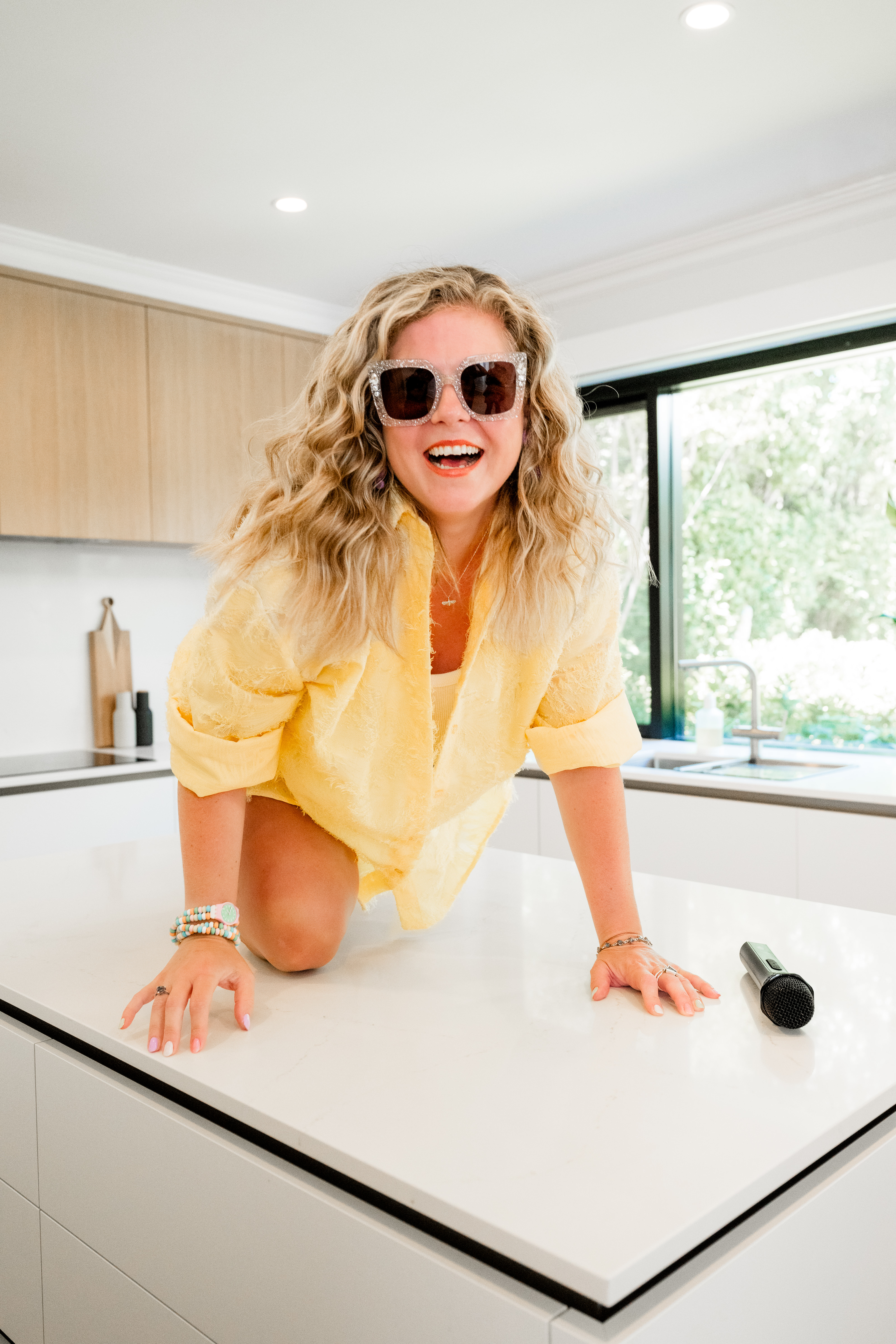 Zoë Condon leaning across a white kitchen island, laughing, with a microphone on the bench beside her