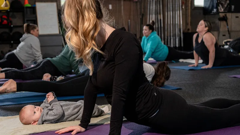 A coach helping a new mom train with her baby on the mat