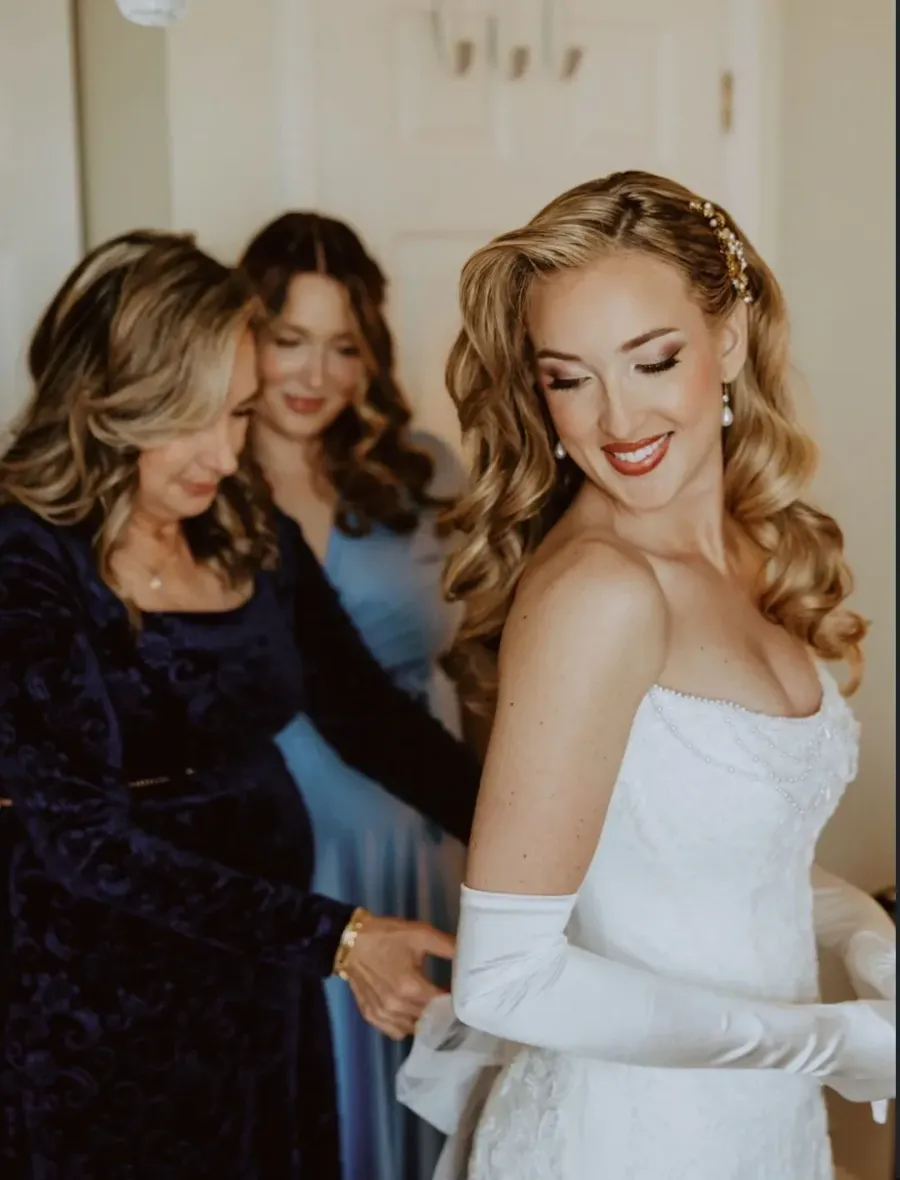 Mother buttoning the back of a bride's dress, both with finished hair and makeup