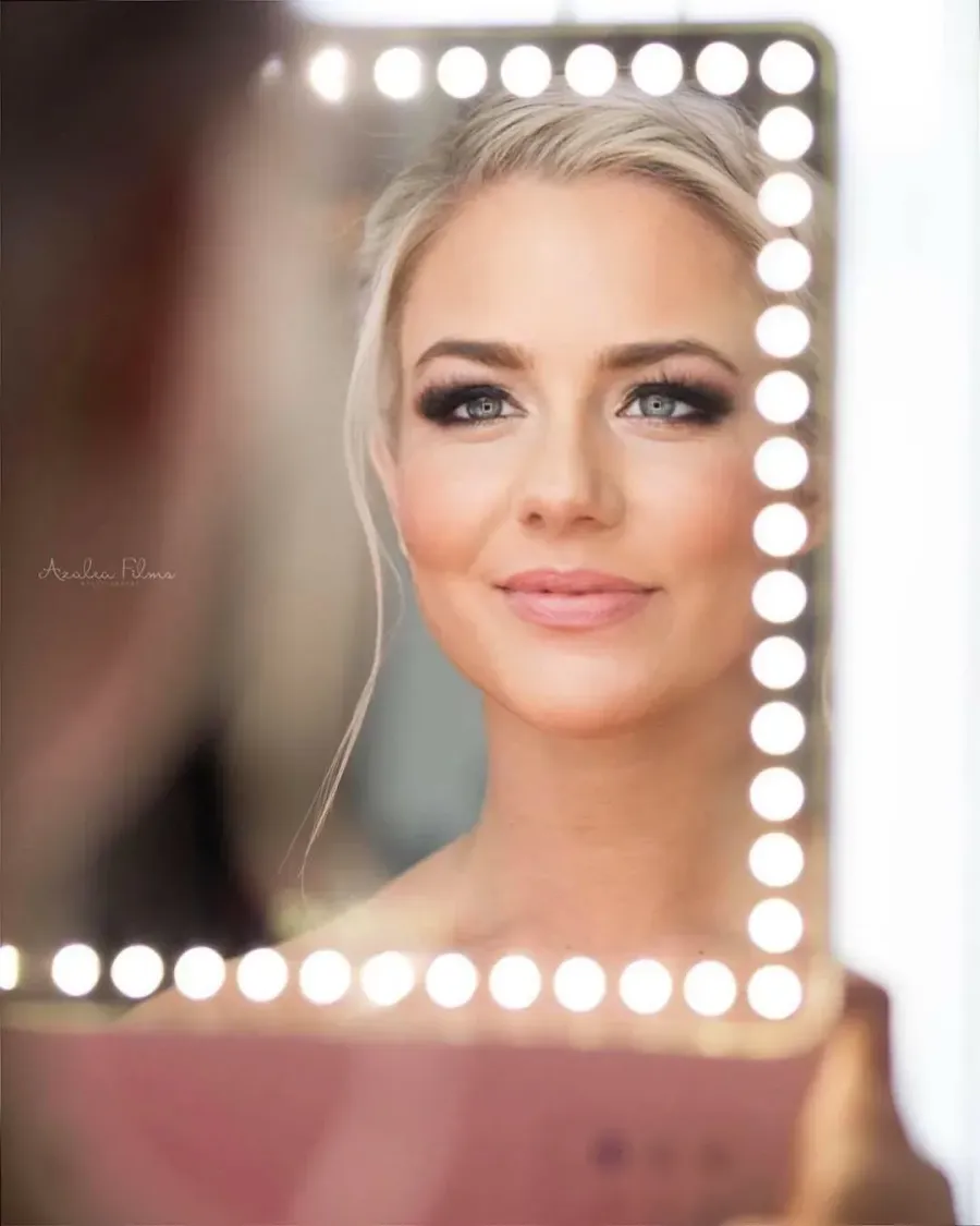 Bride with soft glam makeup and a sleek updo reflected in a lighted vanity mirror