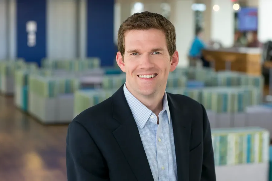 Corporate headshot of a man in a gray blazer and light blue shirt in a modern office