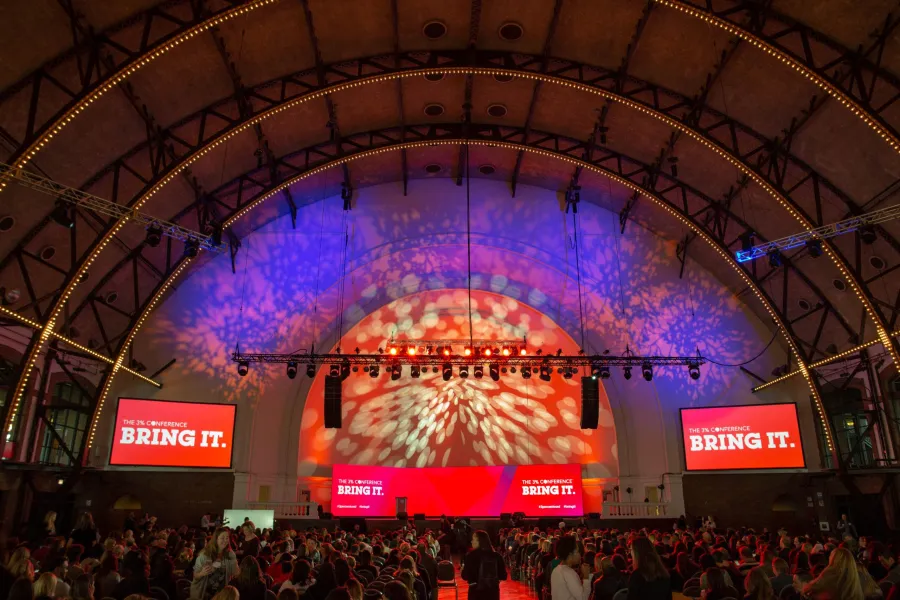 Conference audience facing a stage lit in red and purple under an arched ceiling