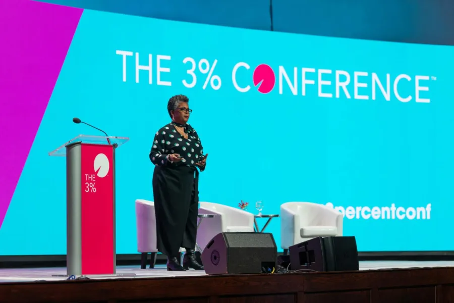Speaker presenting on stage in front of The 3% Conference screen