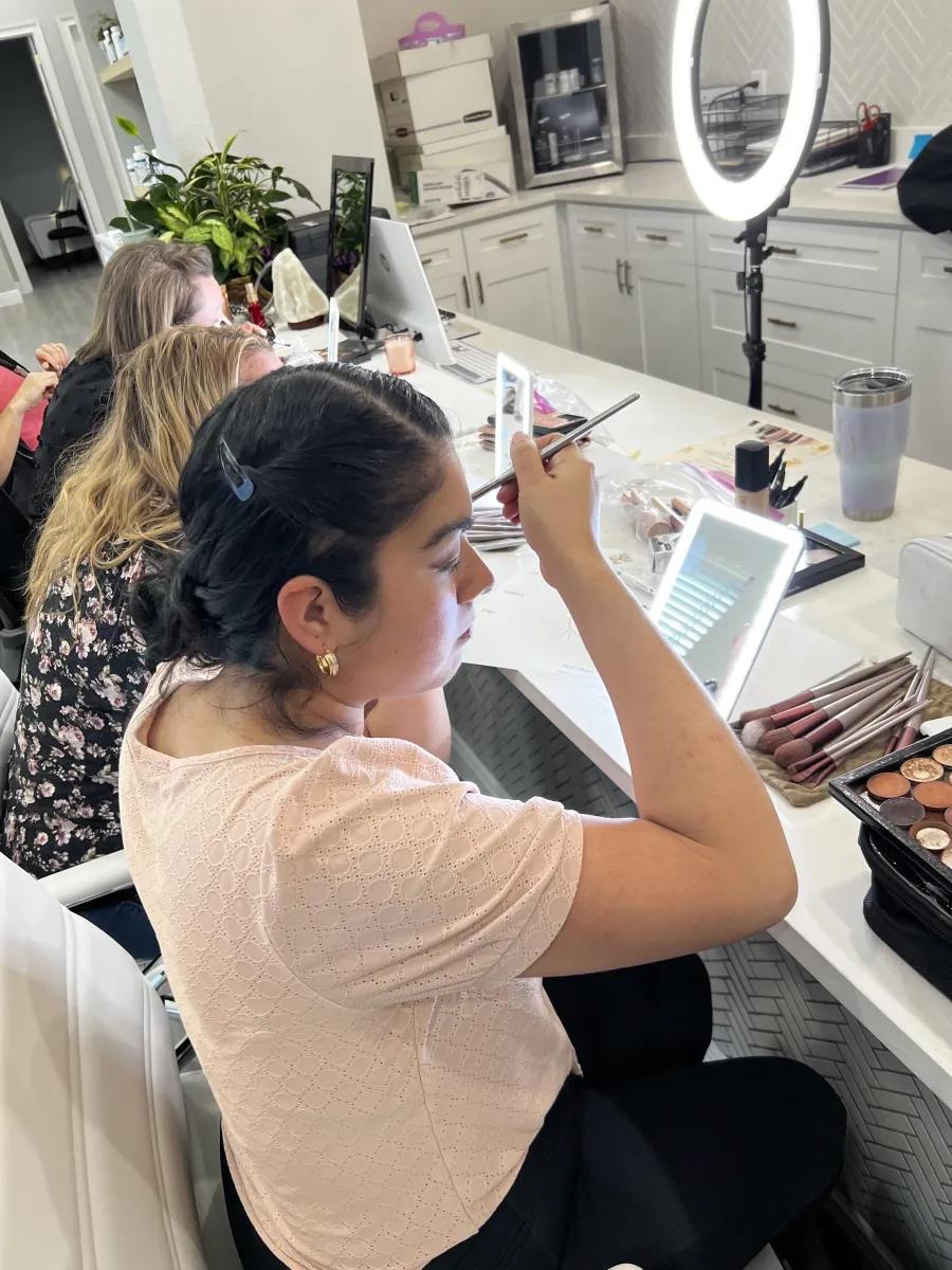 A client applying her own makeup at a lighted mirror while Paula guides her