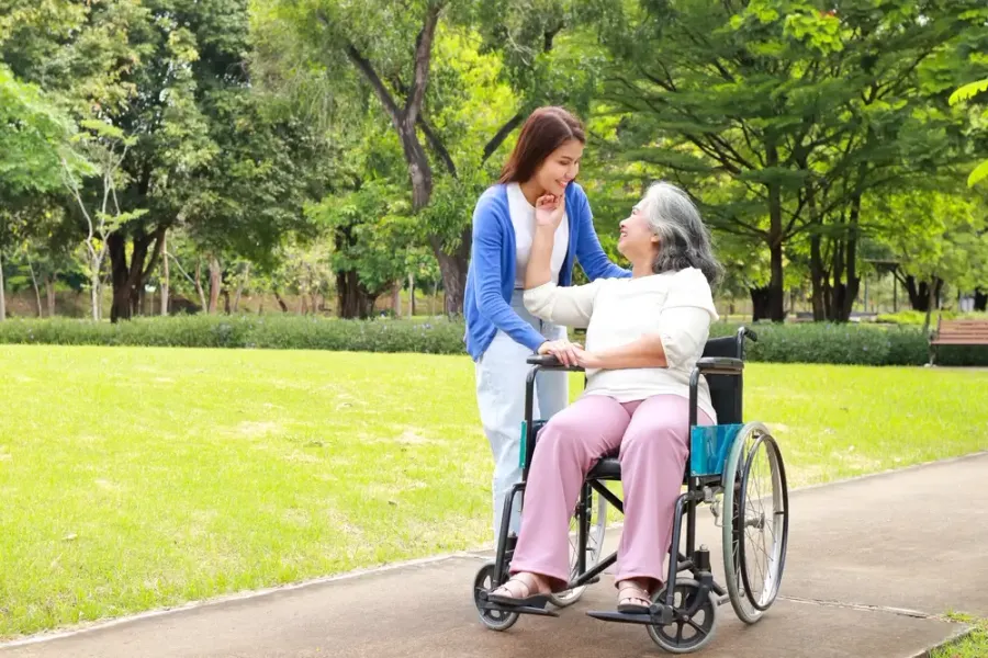 Caregiver walking beside a senior woman in a wheelchair on a sunny park path