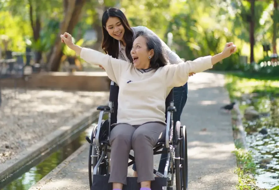 Senior woman in a wheelchair raising her arms outdoors, a caregiver smiling behind her