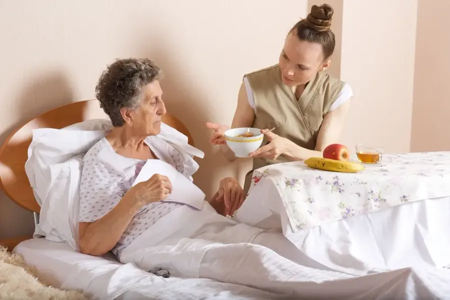 Caregiver bringing a meal tray to a smiling senior woman resting in bed