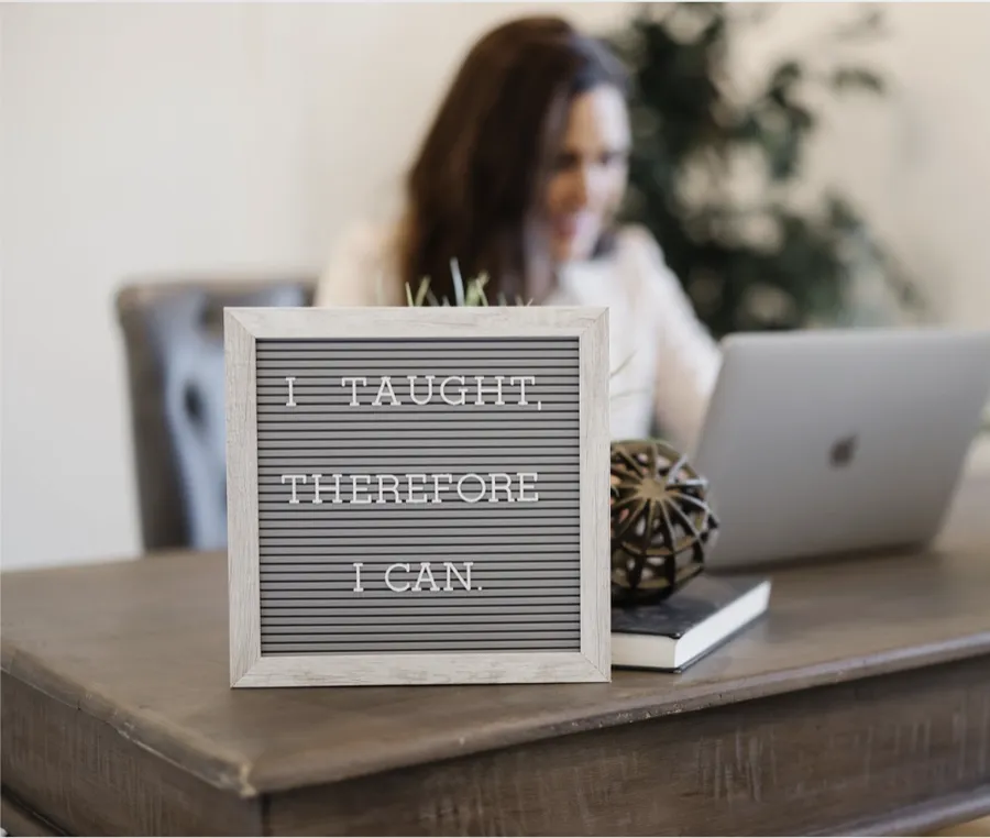 A letter board on a desk reading “I taught, therefore I can” while a former teacher works on her laptop behind it