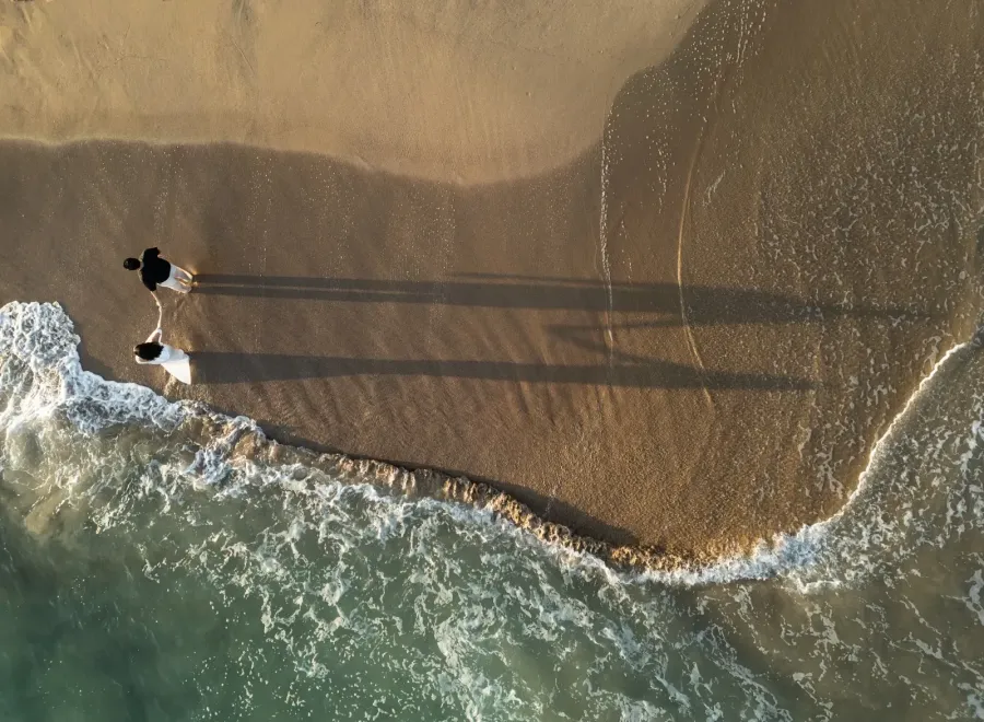 Isamar y Brian caminan de la mano junto a las olas; sus sombras largas se dibujan en la arena