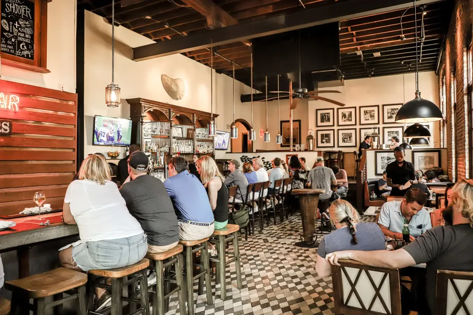 Guests dining and enjoying drinks at the bustling Oyster House bar and restaurant with televisions and framed art in a cozy, lively setting.