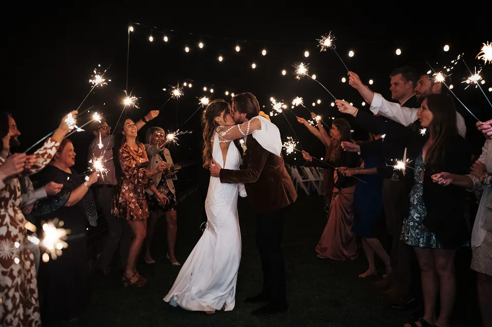 A newly married couple sharing a kiss during a sparkler send-off beneath string lights.