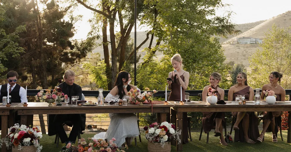 A wedding party listening to a toast at a long outdoor table, with trees and foothills behind them.