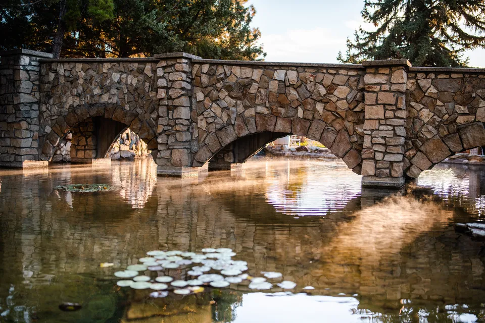 The stone bridge reflected in the estate pond, with lily pads and trees nearby.