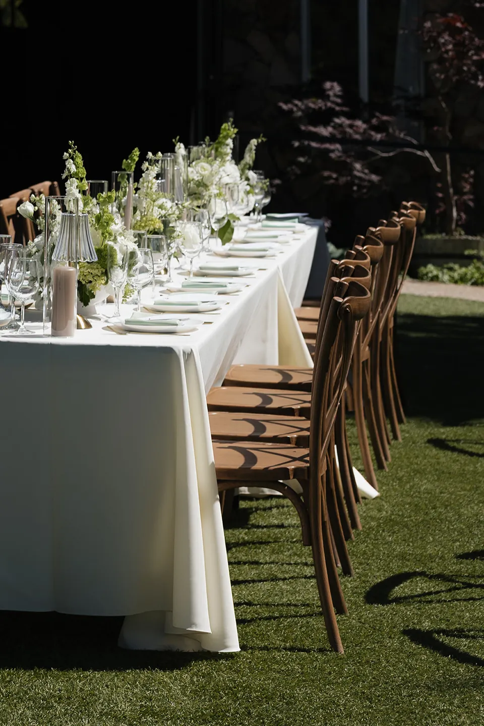 An outdoor dining table set with cream linens, glassware, flowers, and wooden chairs.