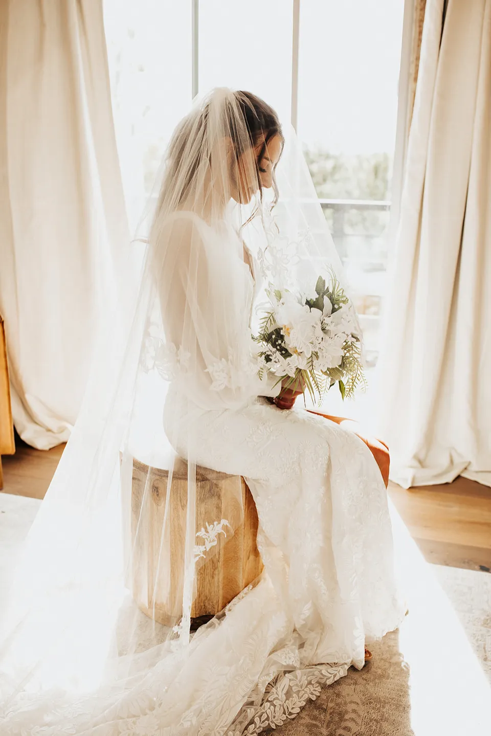 A bride seated beside a sunlit window in the bridal suite, holding her bouquet.