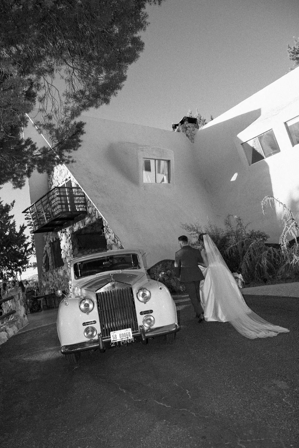 A black-and-white portrait of a newly married couple walking beside a vintage car at the estate.