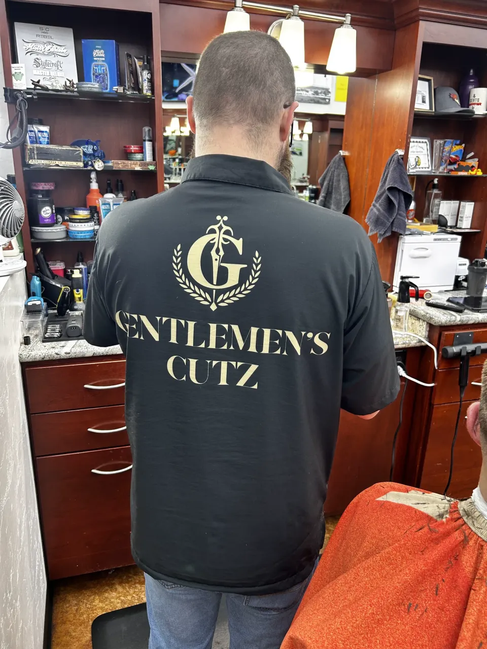A barber's shirt with the gold Gentlemen's Cutz crest and wordmark on the back