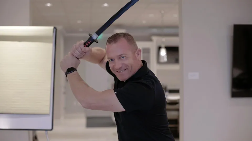 Gene Slade raises a katana over his shoulder in a bright training room, smiling at the camera
