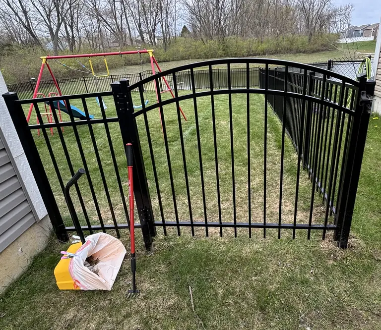 Black arched backyard gate with cleanup tools and a green play yard beyond.