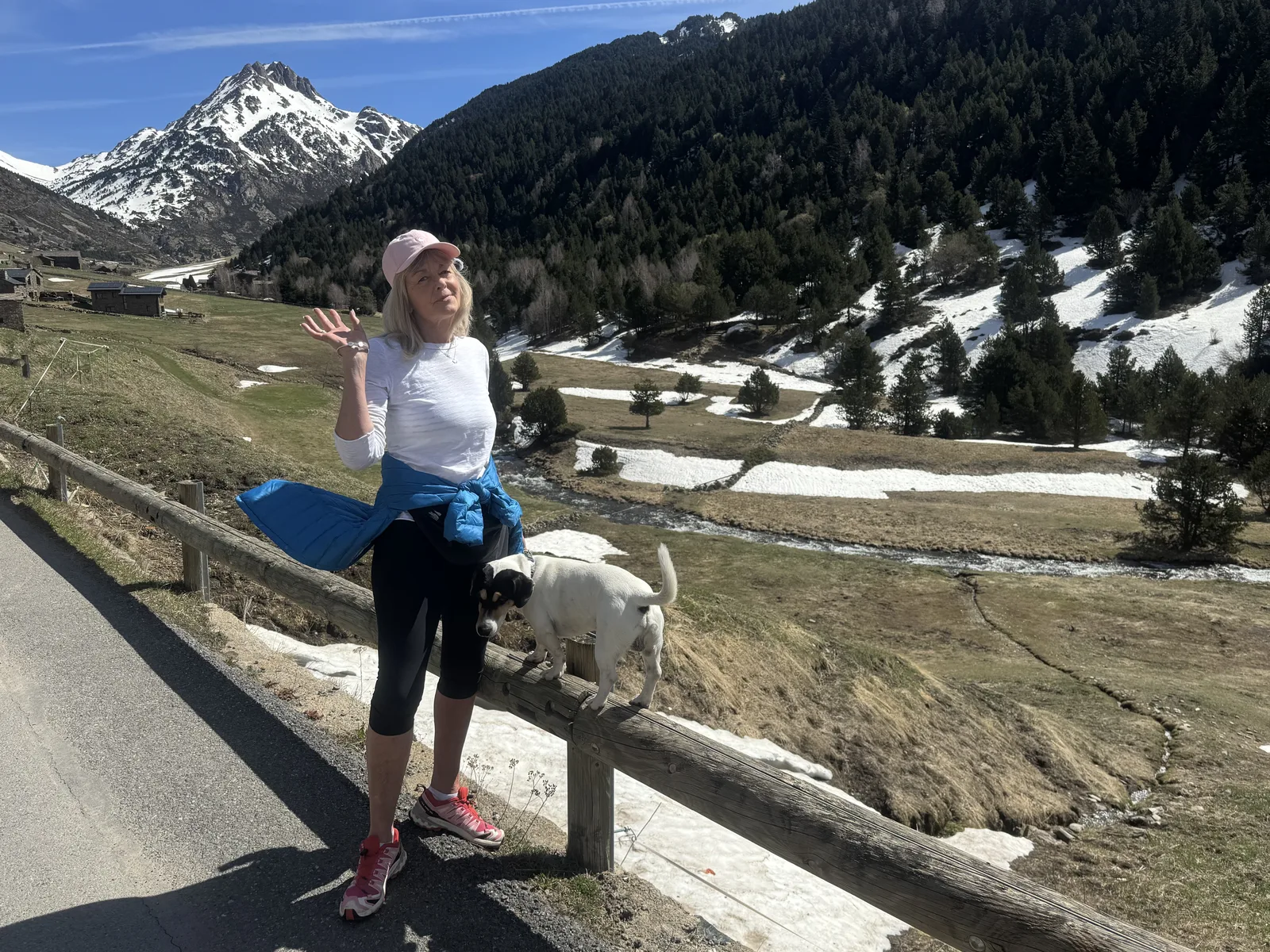 Emma waving on a mountain road in Andorra with her dog Mikey standing on the fence rail, snow-capped peaks behind