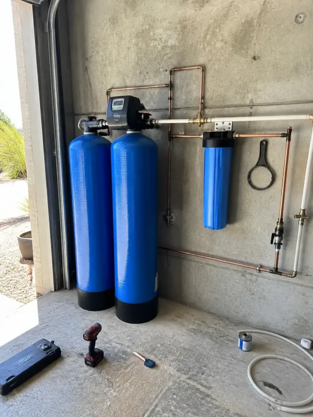 Whole house water filtration system with blue tanks and copper pipes installed in a garage in Mesa, AZ.