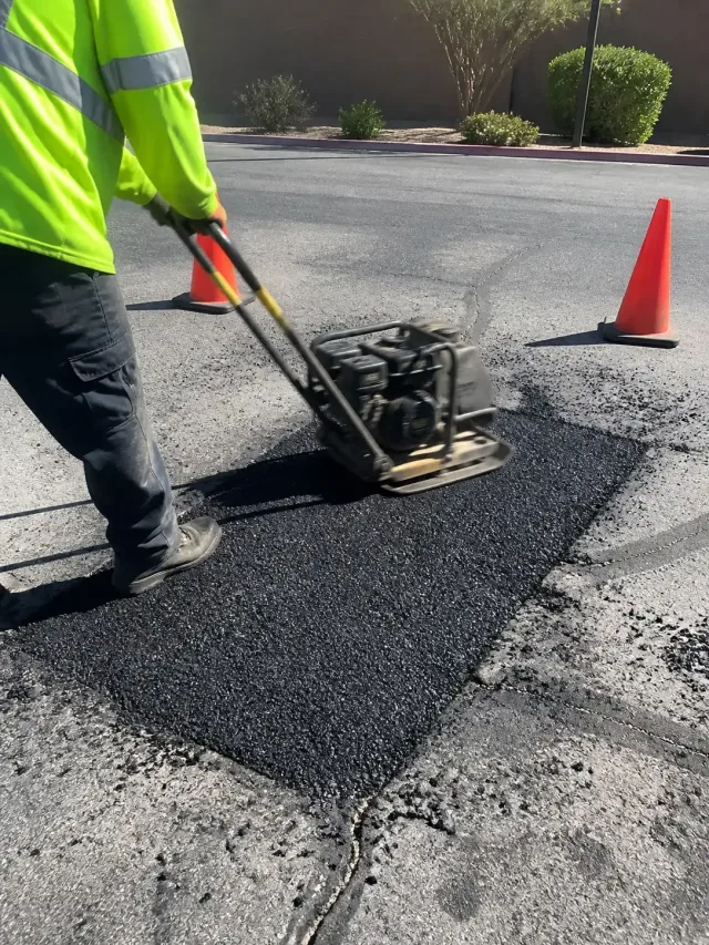 Technician using a vibrating plate compactor on a fresh asphalt patch in a Scottsdale, AZ business lot.