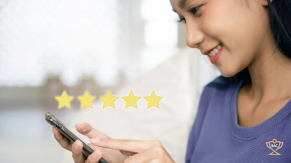Young lady smiling as she's checking her business' customer reviews on her mobile phone, with 5 gold stars elevated above the phone.