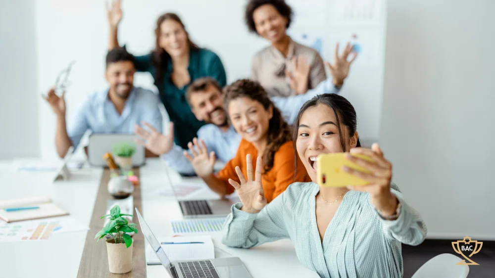 People seated at a work table smiling and waving at mobile camera phone being held by lady taking a selfie in front of the group