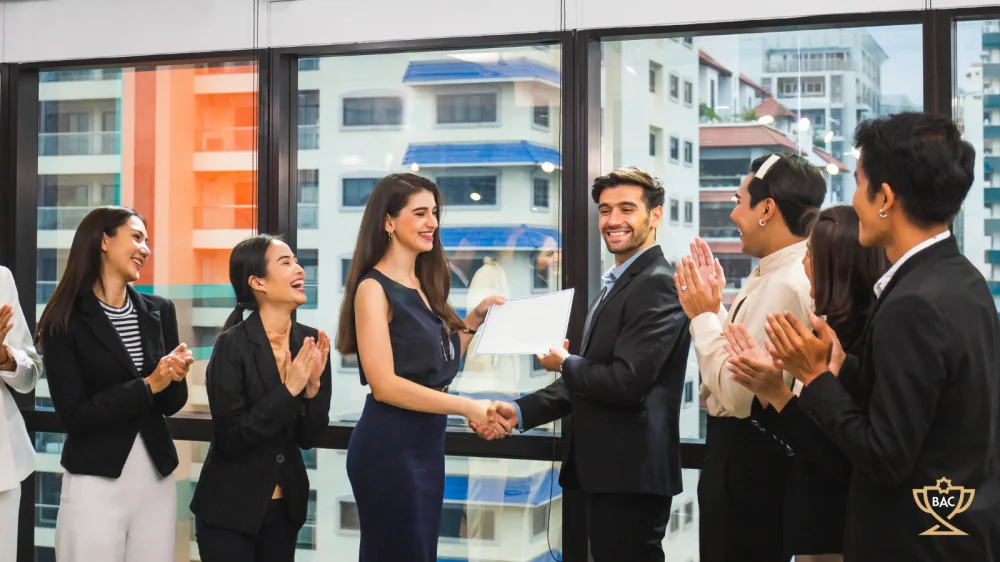 Man in suit receiving certificate of achievement, whilst being applauded by surrounding colleagues.