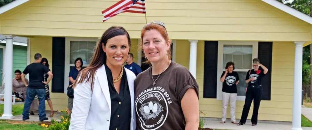 Two women standing in front of a newly completed home with an American flag, representing community partnership and affordable housing support through Fuller Center for Housing of Delaware.