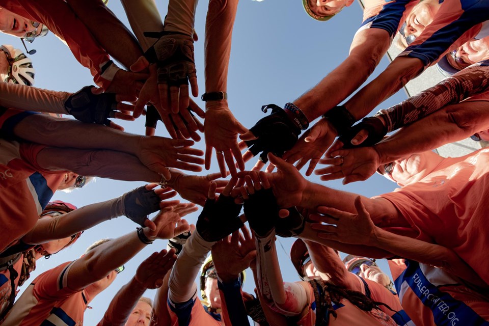 Group of volunteers reaching hands together in unity against a blue sky, symbolizing teamwork and community partnership for affordable housing.