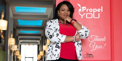 Coach Tamara, an Executive Career, Brand & Business Coach and founder of iPropel You, stands smiling in a professional hallway in front of a bright red door. She wears a red top, black pants, red high heels, and a stylish black and white floral blazer.