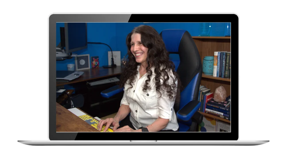 Victoria Essner, seated at her assistive technology workspace, smiling confidently with a braille keyboard in front of her and a large screen behind.