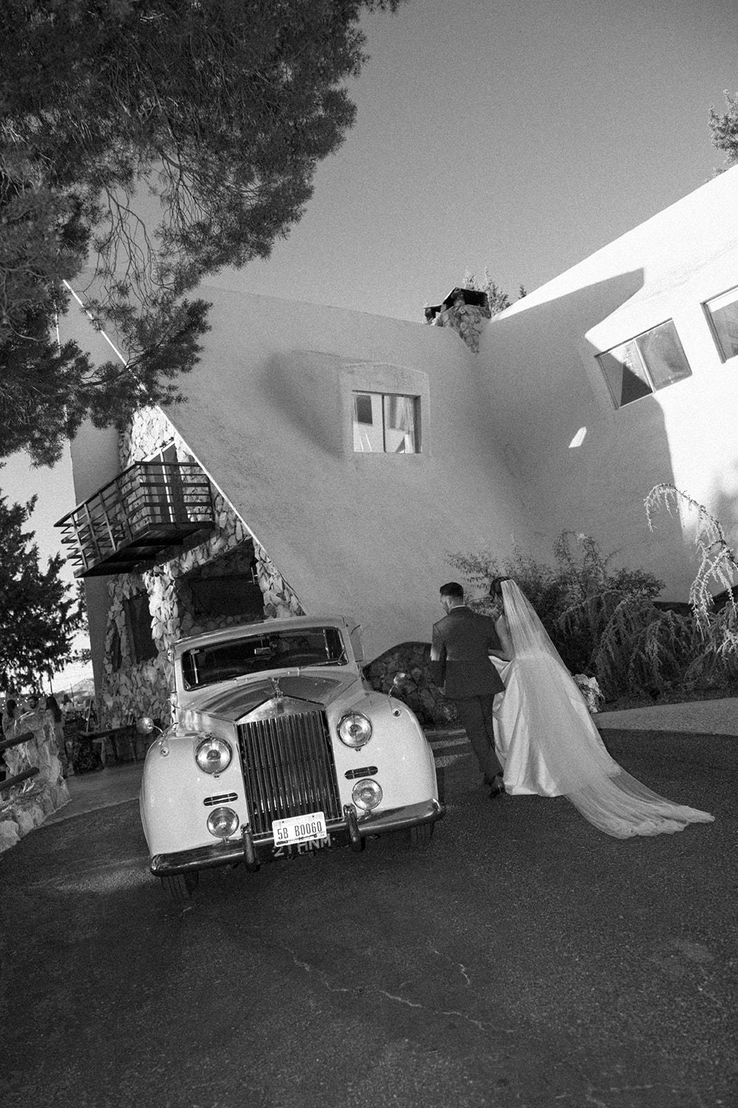 Black-and-white wedding portrait beside a vintage car and the stone estate