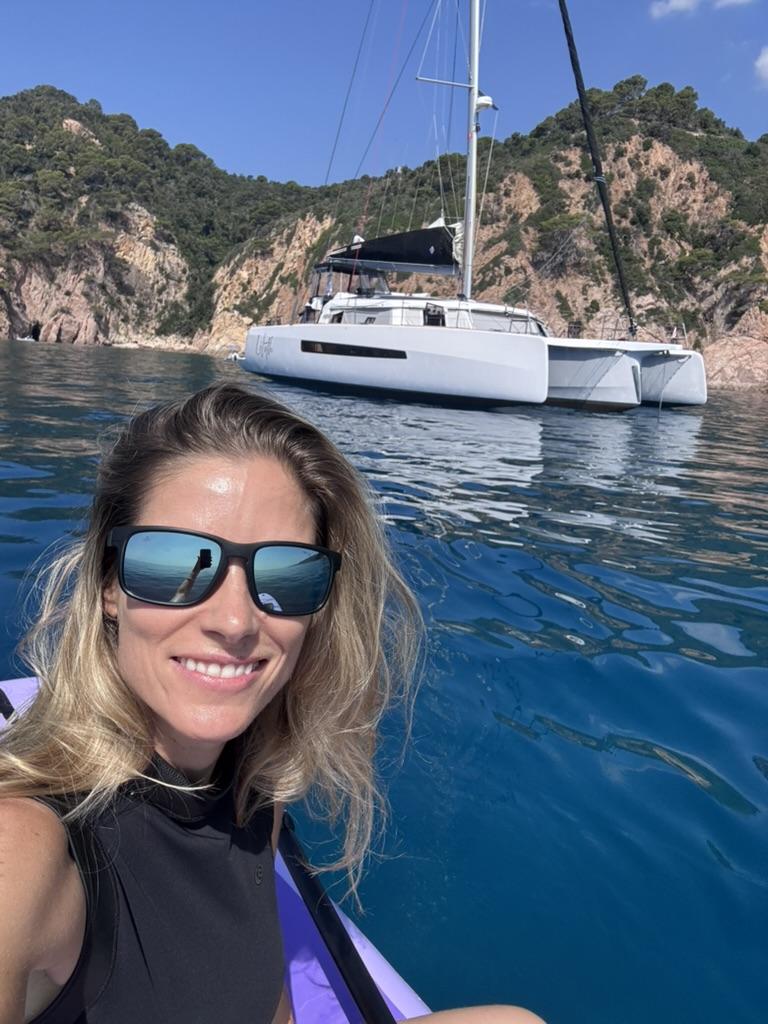 Fanny in the sea, smiling, with the trimaran Lilith anchored behind her below green cliffs