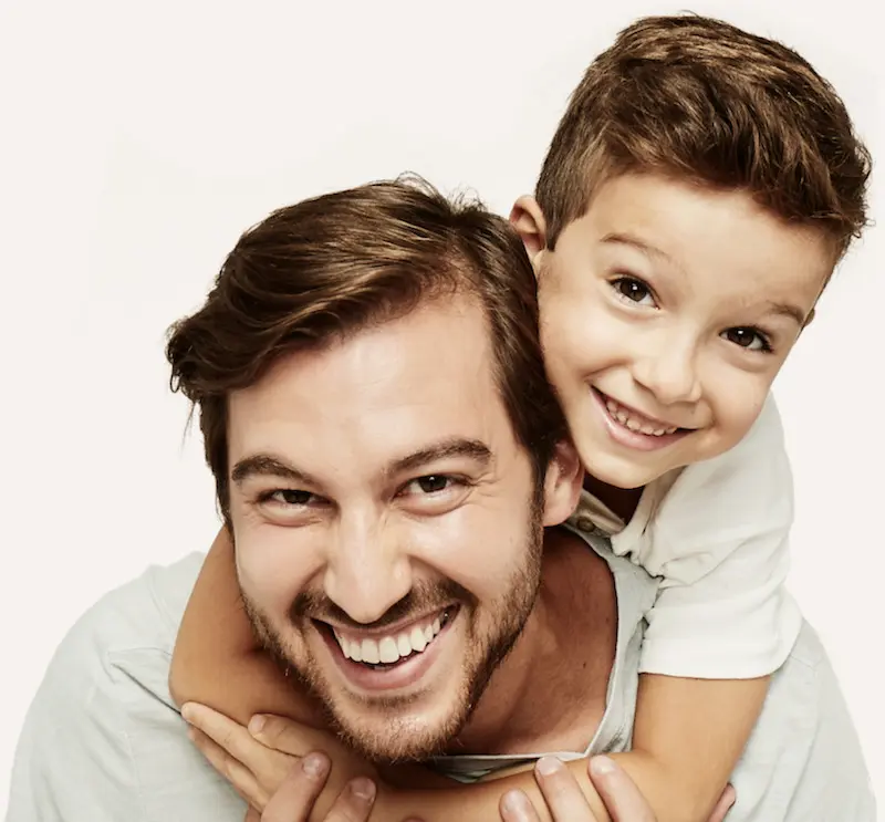 Padre latino sonriendo mientras su hijo lo abraza por la espalda