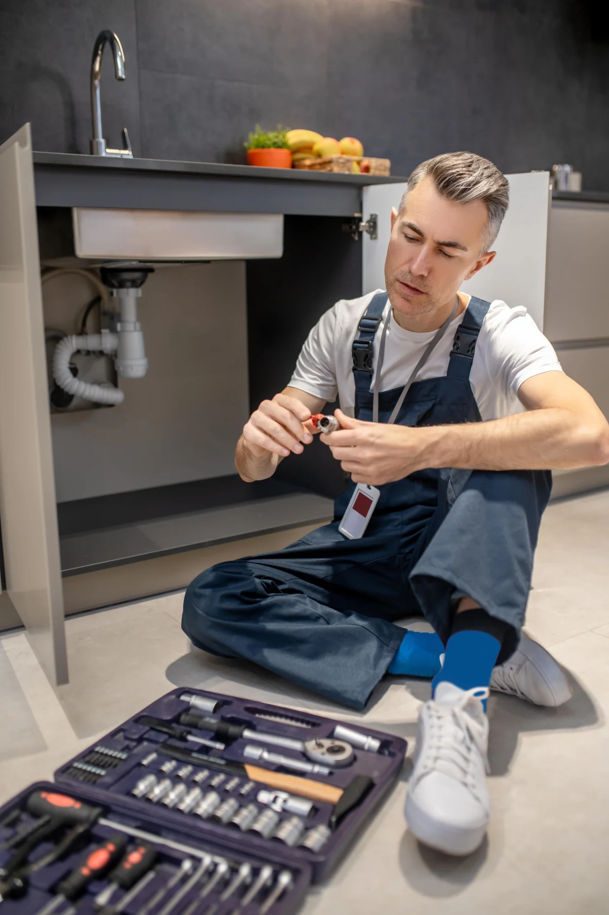 Plumber under a sink with full toolkit, diagnosing a hidden leak