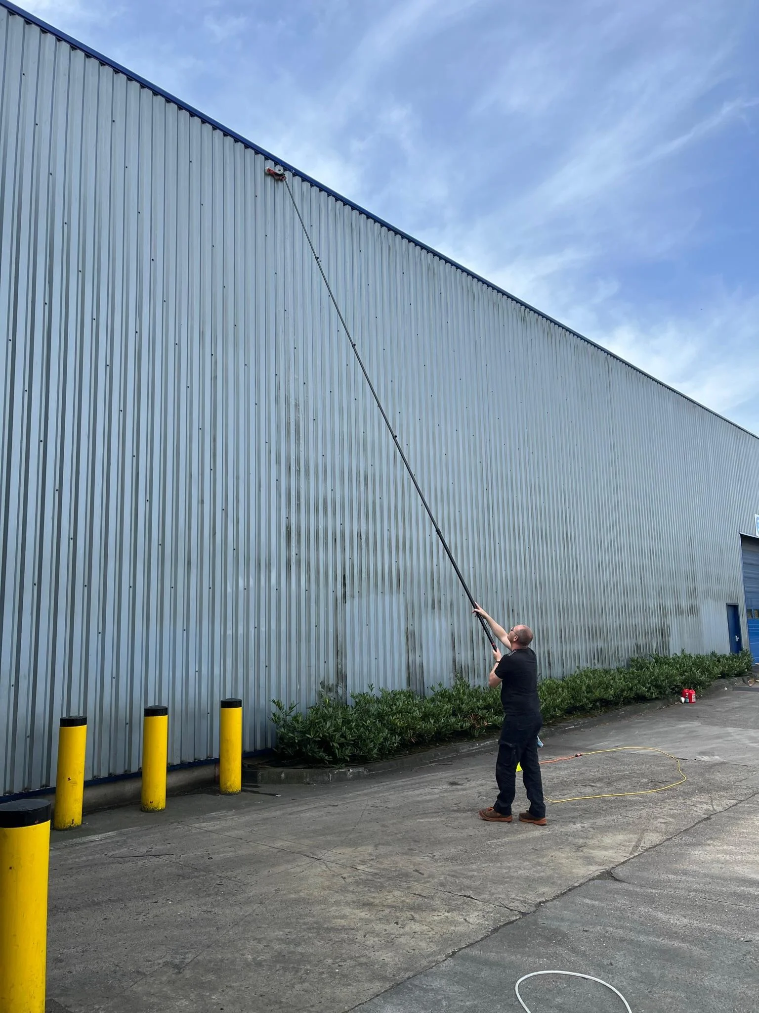 Technicien Surfaclean nettoyant le bardage d'un entrepôt industriel à la perche carbone, depuis le sol, sans nacelle