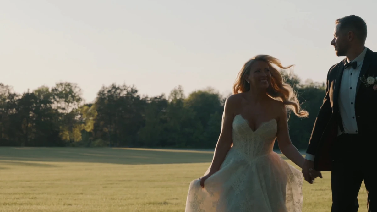 Still from a highlight film: the bride and groom walking hand in hand through a field at sunset
