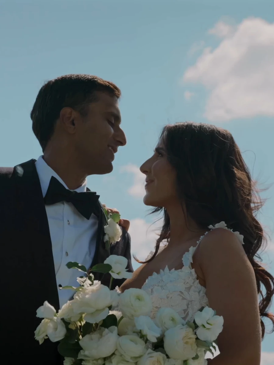A bride and groom smiling at each other under a blue sky, her bouquet in hand
